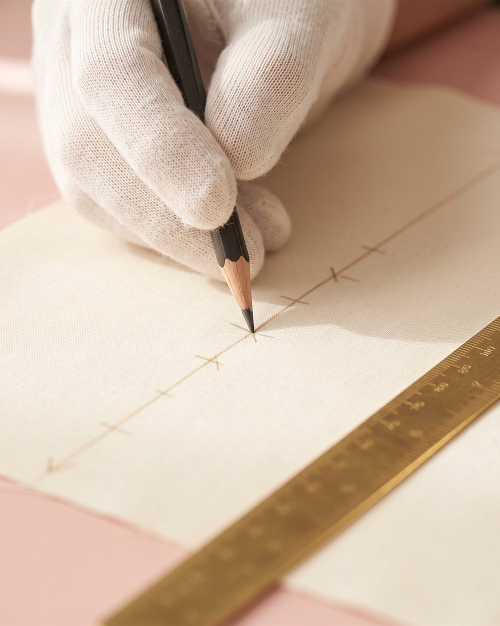 A gloved hand marking a cutting line on satin with a pencil and a brass ruler